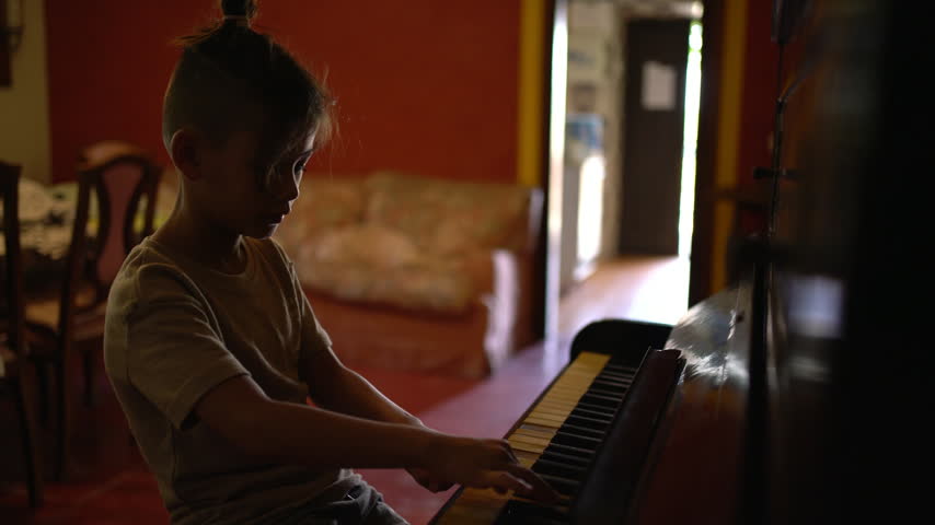 A boy plays piano in silhouette beside an open doorway in a rustic home living room in Tarragona, Spain