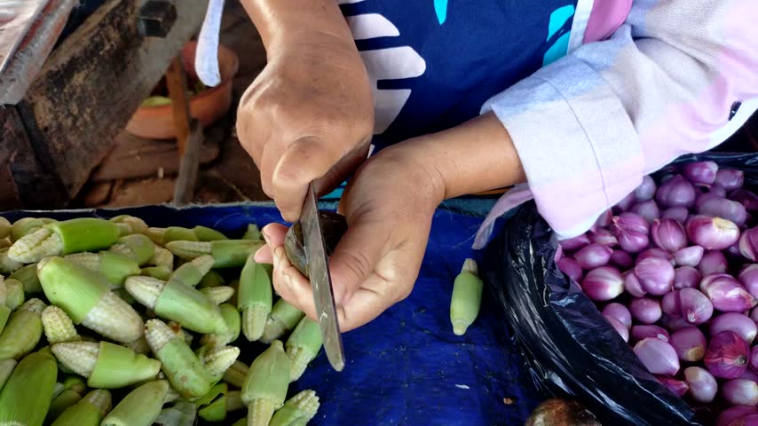 A close-up shot of a market vendor peeling *jengkol* beans and extracting the kernels, with other vegetables being sold in the background.