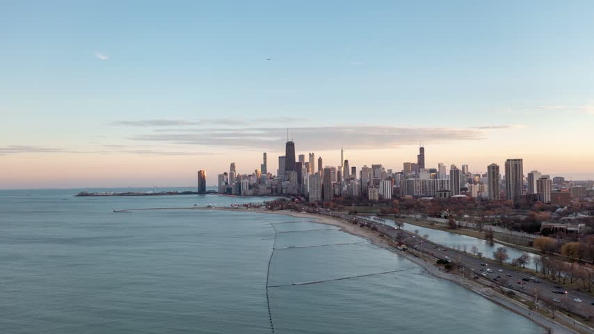 Wide aerial hyperlapse overlooking Chicago’s waterfront, where Lake Michigan meets a curving sandy beach, parkland and busy roads below the distant skyline and pastel sky.