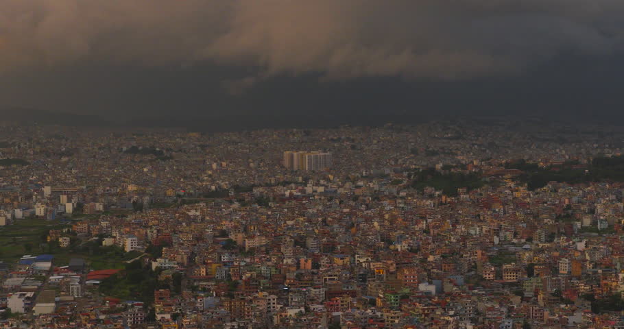 Kathmandu, Nepal, wide drone shot over the dense urban concrete sprawl of Kathmandu Valley highlighted by warm sunset golden hour glow beneath dark monsoon storm clouds.