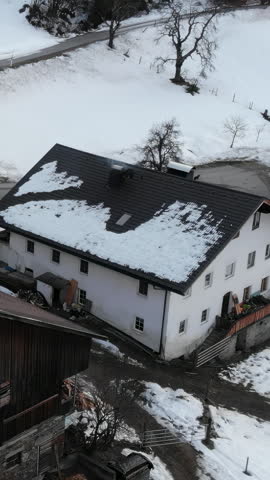 High oblique aerial of rural farmhouse roof with dark metal roofing, melting snow patches, and wet slush runoff during winter thaw on snowy hillside under overcast sky