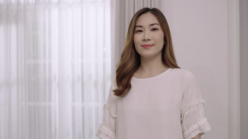 Happy and confident, beautiful, Asian woman with long brown hair standing smiling with arms crossed while looking at camera in living room at home. Closeup attractive young female with a Siam smile. 