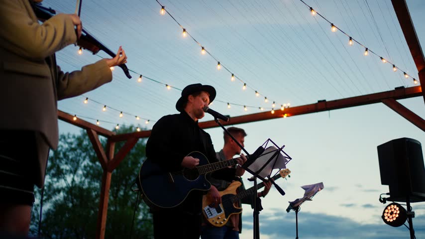 Outdoor evening band performance under string lights with guitarist singer bassist and violinist playing live music on wooden stage at sunset