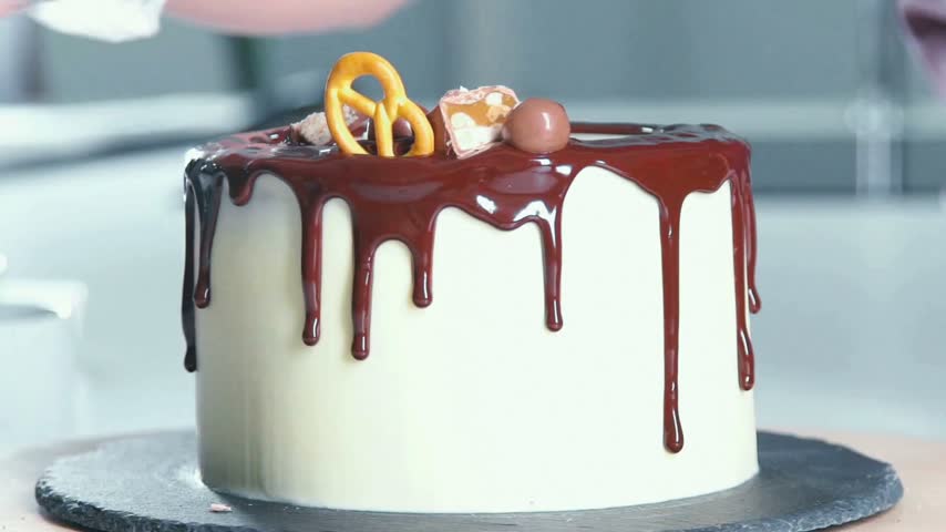 A baker skillfully decorates a white cake with rich chocolate and various toppings, including candies and pretzels, creating an eye-catching dessert perfect for celebrations.