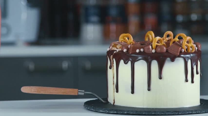Baker slices a chocolate cake topped with ganache, chocolate pieces, and pretzels in a modern kitchen setting. Kitchen utensils and ingredients visible in background.