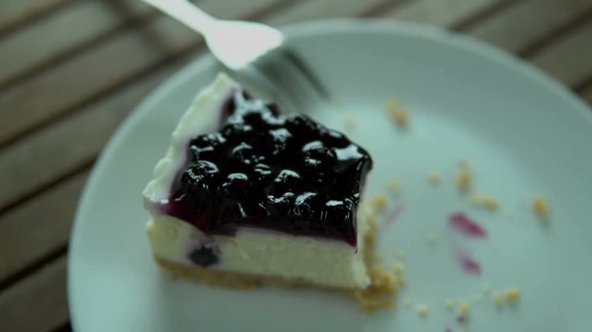 Slice of blueberry cheesecake placed on white plate. Fork rests beside cake. Background features wooden table surface, creating a simple and inviting dessert display.