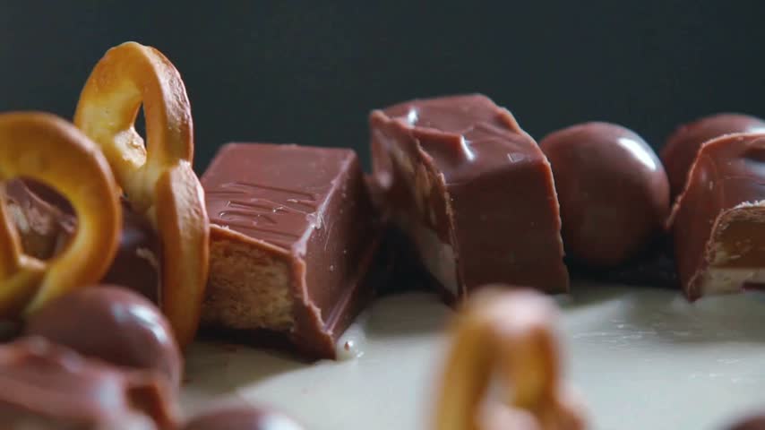 Chocolate bars and pretzels lay together on a white surface as thick sauce drizzles over them, highlighting creamy texture and contrasting shapes, all in a well-lit setting