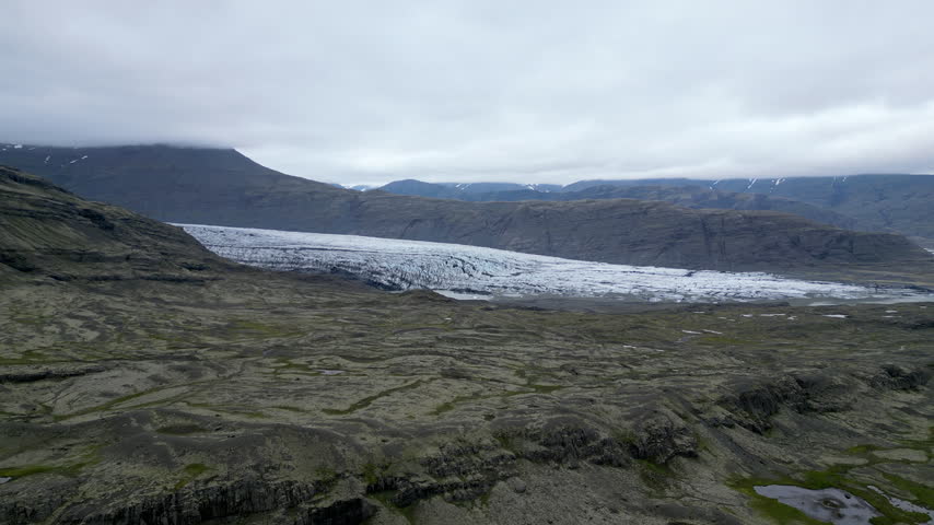 A majestic Skálafellsjökull glacier stretches across a rugged Icelandic mountain valley under a cloudy sky, creating a serene and epic natural atmosphere.