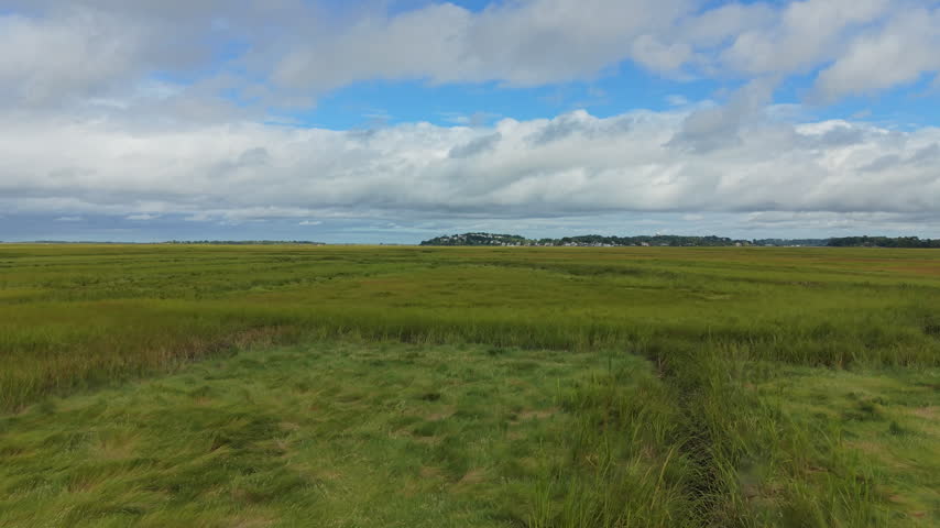 Low aerial flight over salt marsh grass with big cloudy sky and distant coastal village, Ipswich Massachusetts, salt marsh
