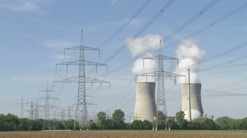 Power lines and transmission towers extend across landscape near nuclear power plant with cooling towers releasing steam, showcasing energy infrastructure.