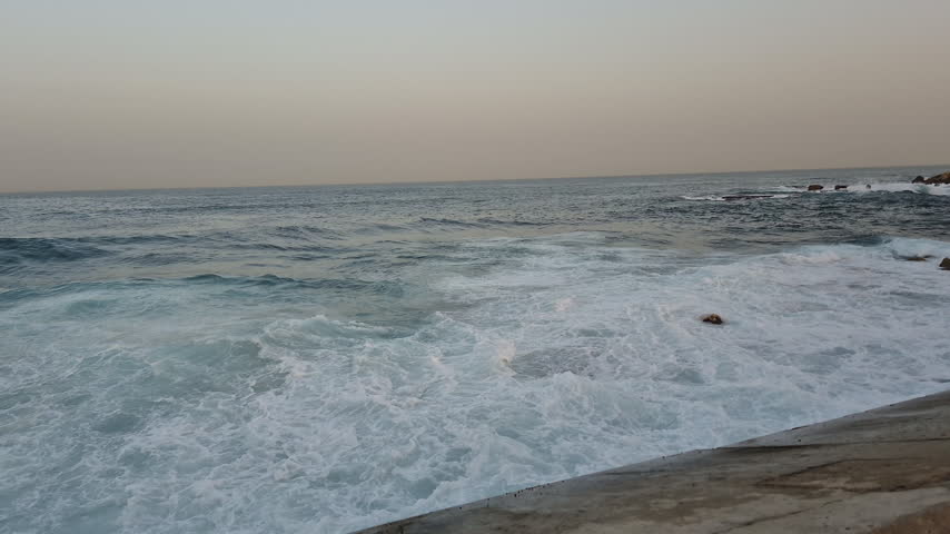 Foaming sea waves break and splash over rocky shallows near the concrete seawall of historic Jaffa Port along the Mediterranean coast under an overcast daytime sky Pan Shot
