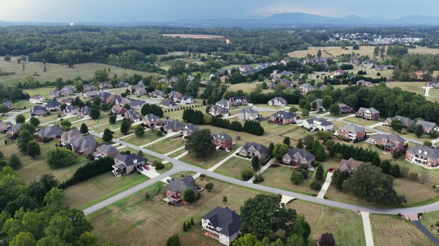 Rooftops of a large lot subdivision spread across rolling Virginia countryside with the Blue Ridge mountains rising faint along the horizon. Aerial rising wide shot