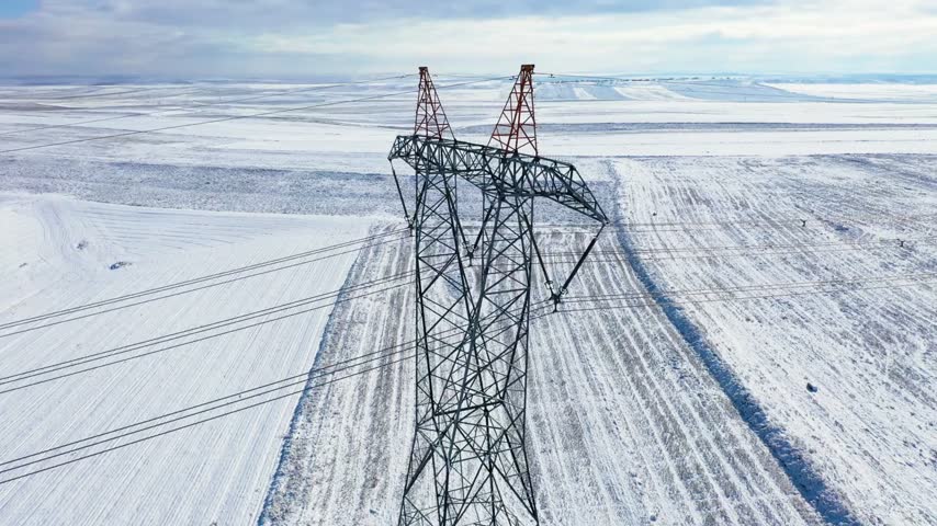 Metal power transmission tower stands tall amidst snow covered landscape. High voltage lines stretch across Winter fields under a cloudy sky.
