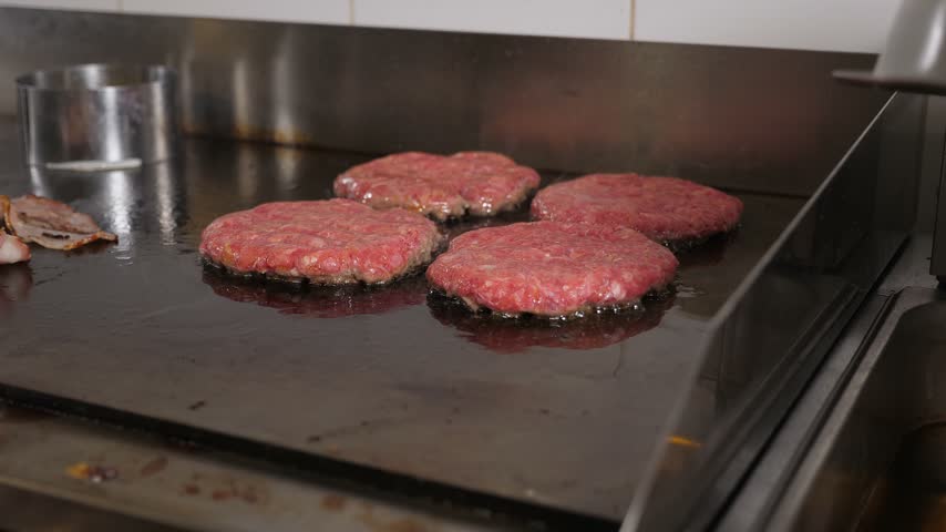 Close-up of bacon and burgers patties fried on a large stove in the restaurant kitchen. The process of cooking juicy cutlets and bacon for burgers.