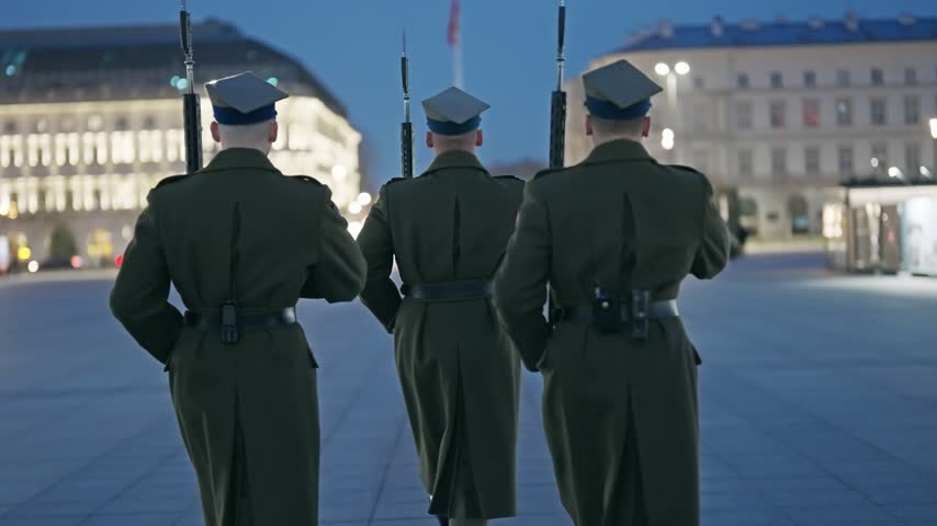 Three uniformed soldiers perform the solemn change of guard ceremony at the Tomb of the Unknown Soldier in Warsaw, Poland. A poignant display of national honor and tradition against the backdrop of an illuminated city at dusk.