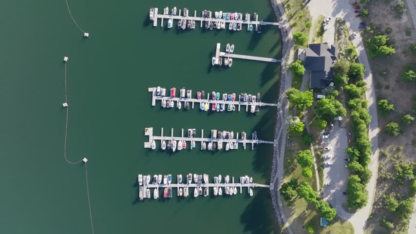 Aerial view of a boat marina on a quiet reservoir during a sunny summer day showcasing anchored vessels wooden piers and coastal park land with parked vehicles and green trees