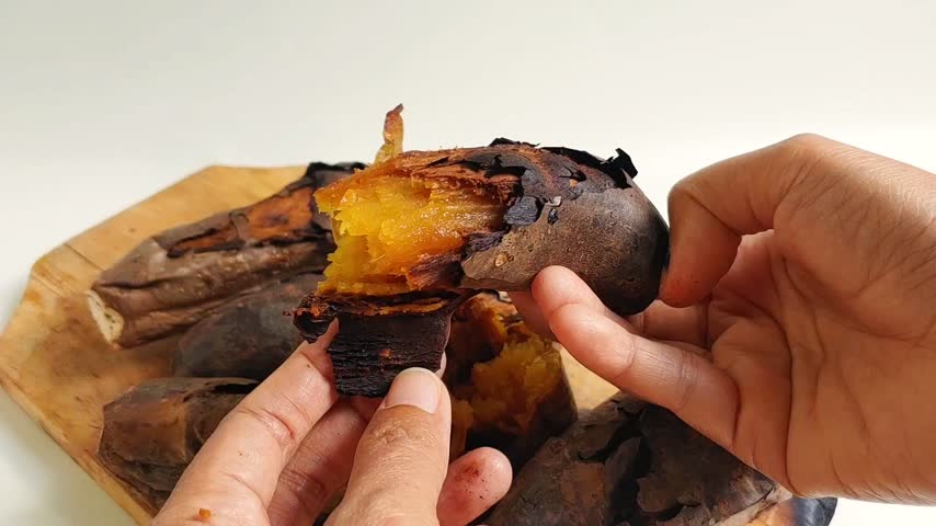 Close-up of a person peeling the charred skin and breaking open a freshly baked, steaming yellow cilembu sweet potato on a rustic wooden cutting board