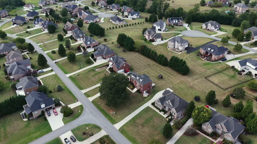 A curving residential street winds between large brick homes on wide green lots and long driveways in an established Forest, Virginia neighborhood. Aerial backwards shot