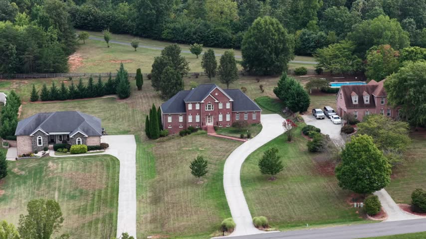Two large brick houses share long curving driveways across open mown lawns backed by dense summer woodland in Forest, Virginia. Aerial forward shot