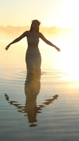 Beautiful young woman in white dress wearing flower crown walking in lake water at foggy golden hour sunrise, throwing water drops and enjoying nature freedom in summer morning.