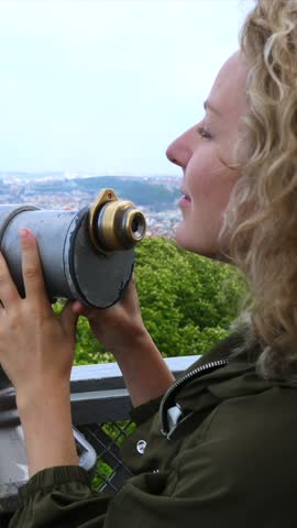 Woman with curly blonde hair looks through viewing telescope above hillside city panorama. Her fingers grip metal housing as she adjusts her gaze toward distant rooftops and hills.