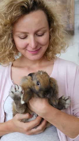 Woman cuddles baby rabbit in gentle embrace. She smiles softly while holding small brown bunny close to chest, creating tender home portrait with calm warmth and careful hands. Curly blonde hair