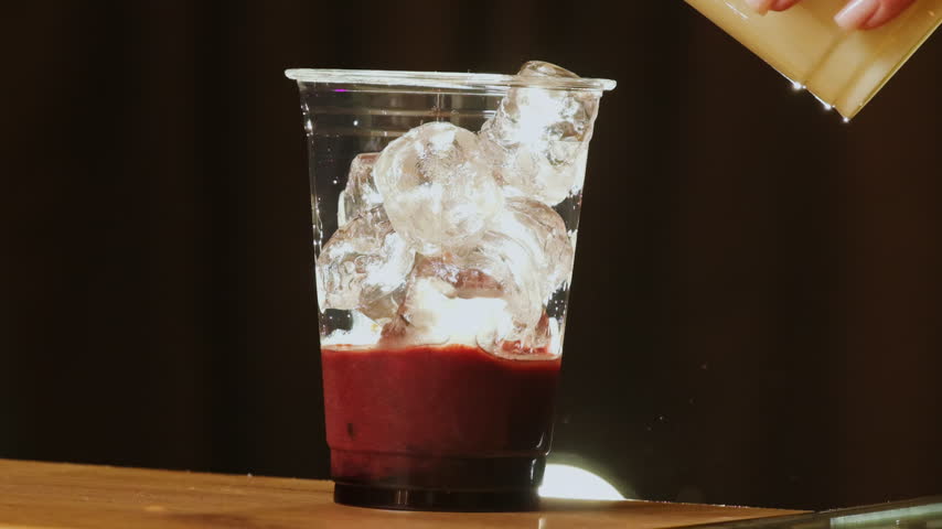 Barista pours syrup over ice and cherry base in clear cup. Woman creates sweet layer as cold berry drink gains glossy texture at cafe bar