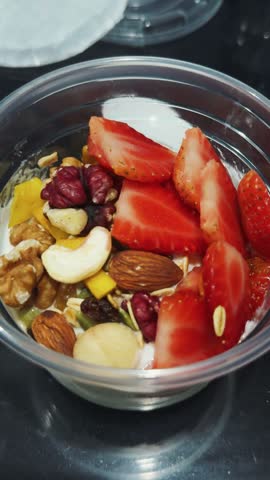 Healthy breakfast featuring a clear plastic cup filled with yogurt, sliced strawberries, various nuts including cashews, walnuts, and almonds, and granola, promoting a balanced and nutritious diet