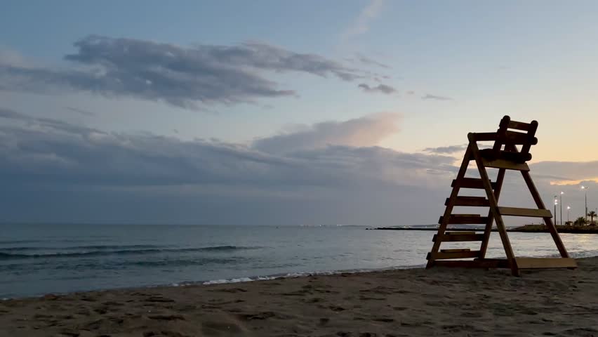 A wooden lifeguard tower stands on a sandy beach on the warm coast of Spain at sunset, with the sea and evening sky in the background. The video begins with the empty rescue tower and later shows the silhouettes of a couple seen from behind against the sea and glowing sunset.