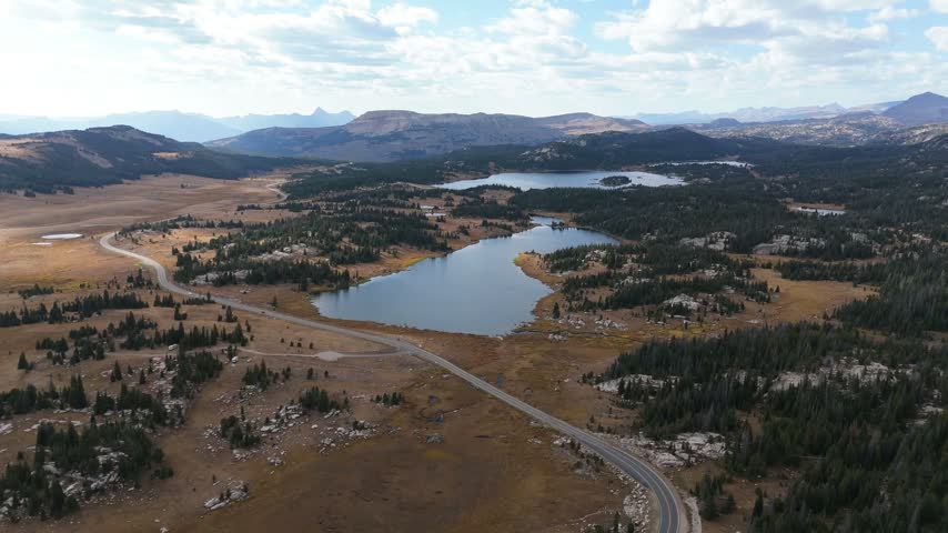Aerial view of Beartooth lake and highway surrounded by mountain peaks and forest under a blue sky, Scenic drone shot of alpine plateau wilderness landscape