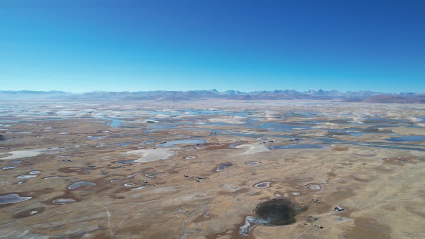 Sweeping aerial view of the golden sand dunes in Zhongba, Tibet, where a winding river cuts through the desert terrain. In the distance, the majestic snow-capped peaks of the Himalayas rise under a cl