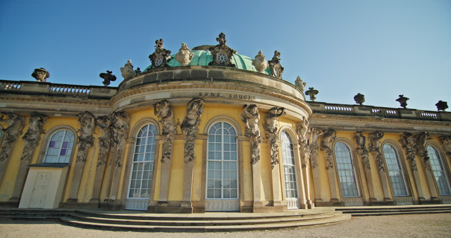 The ornate Rococo exterior of Sanssouci Palace in Potsdam, Germany, has arched windows, carved reliefs, and roof sculptures.