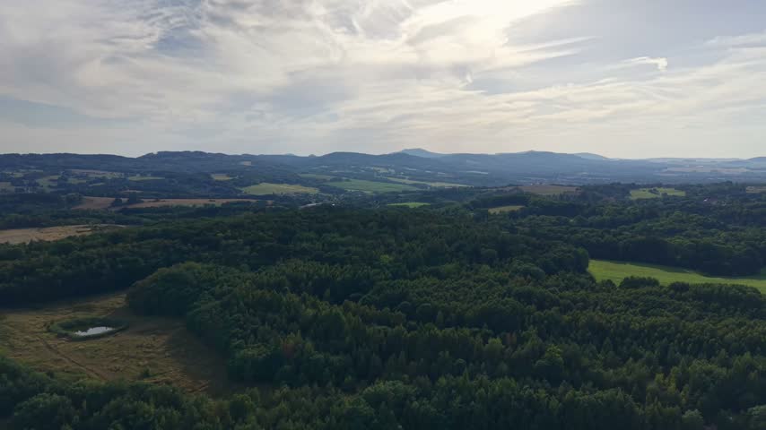 Wide aerial view over patchwork fields weaving through rolling forests, with small pond in foreground, distant village buildings, and bright daylight under scattered clouds
