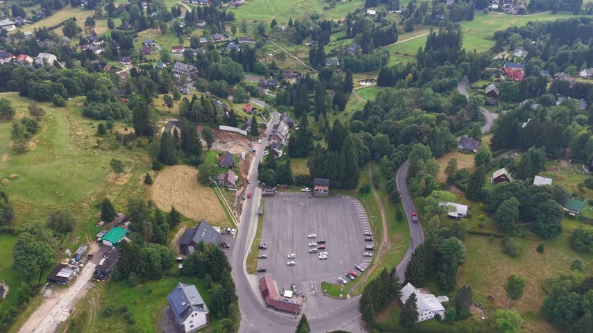 Elevated drone view of rural town with winding roads, scattered houses, and visible construction area beside large parking lot, surrounded by forested hills and open fields