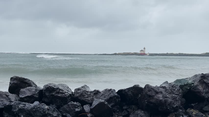 Dramatic footage of stormy sea waves hitting rocky coastline shore with white lighthouse beacon in background during cloudy day.