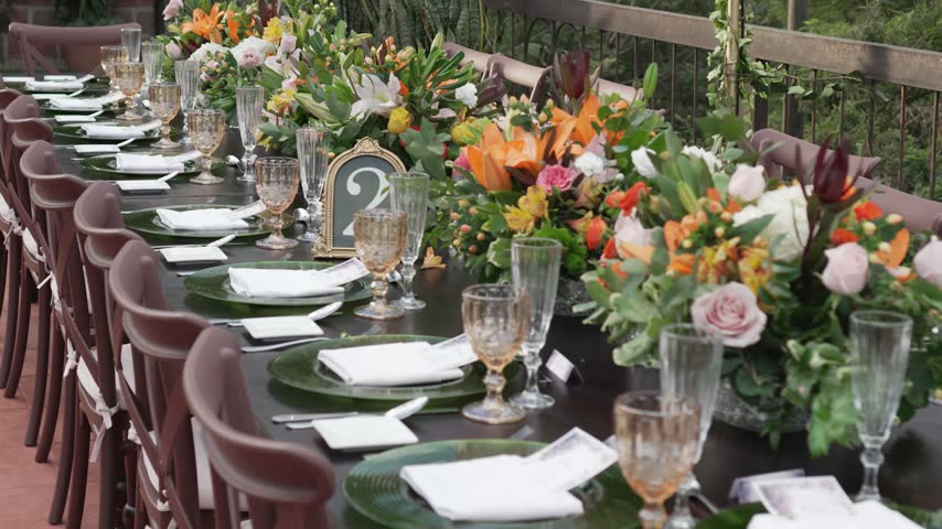 Slow motion footage showing beautifully decorated long dining table for celebratory event featuring lush flower arrangements green plates gold rimmed glasses and numbered decorative frames.