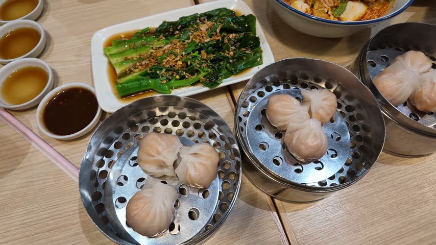 High-angle panning shot of dim sum, bok choy, and noodle soup on a restaurant table.