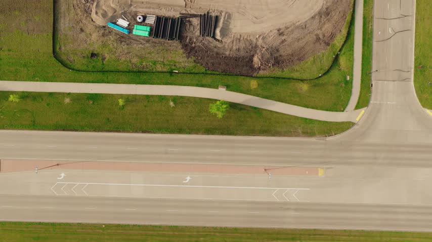 Top down aerial of a construction site in south Fargo, North Dakota: graded sand, stacked concrete pipe, a yellow excavator digging a new basin, work trailers and haul tracks.