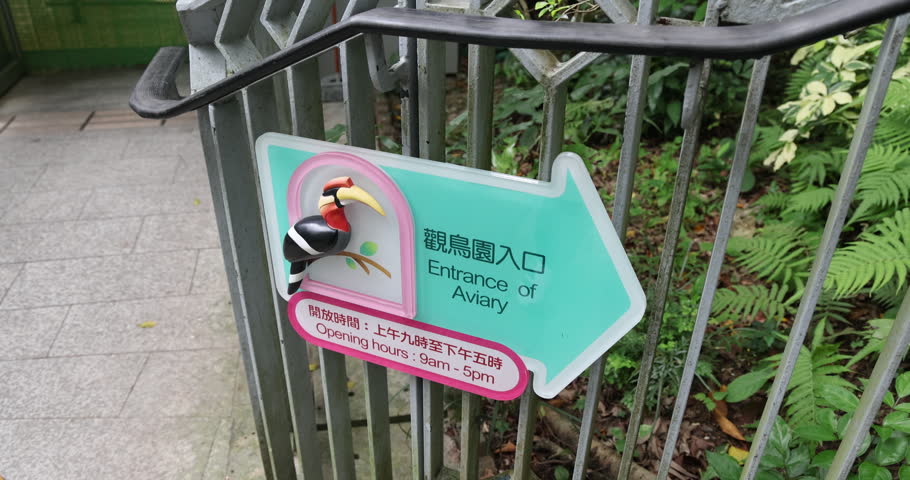 Aviary entrance sign with hornbill illustration attached to metal fence. Bilingual English and Chinese text with opening hours in outdoor park environment, wayfinding signage, nobody.