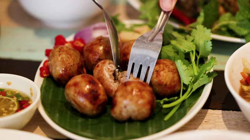 Close-up of person using spoon and fork to cut grilled Thai sausages in Bangkok.