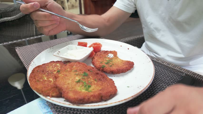 A person enjoys crispy golden potato pancakes with sour cream and sliced tomato at a wicker table in an outdoor café, natural lighting enhancing the appetizing textures and cozy dining atmosphere.