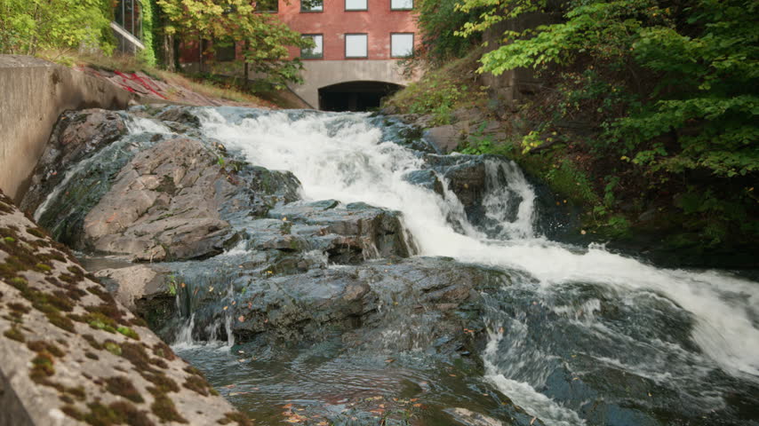 Foamy cascade tumbles over dark rock slabs beneath a red brick building framed by green trees, with fast current churning through the narrow channel.