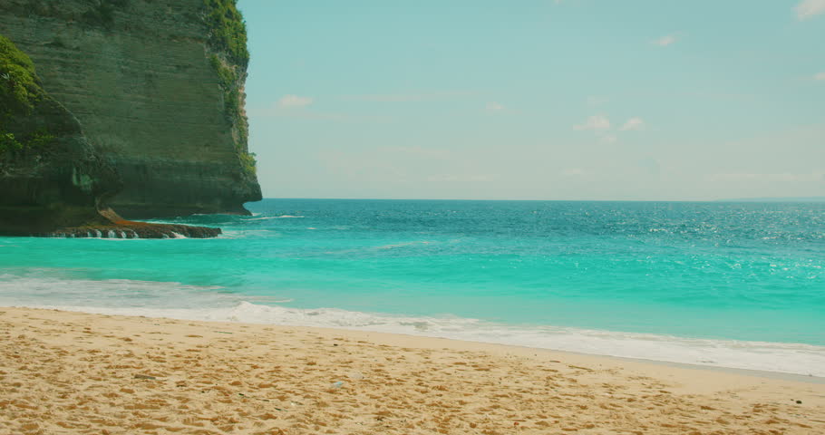 Turquoise ocean waves crash onto white sand beach below green cliff at Kelingking Nusa Penida Bali.