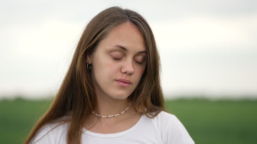 Young woman opens eyes in field. Wind blows long brown hair. Portrait highlights natural female face. Girl reveals calm expression. Woman looks away into green field. Face forms happy big smile.