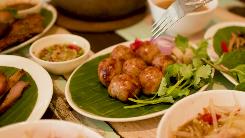 Close-up of hands cutting grilled fermented sausages on a banana leaf in Bangkok, Thailand.