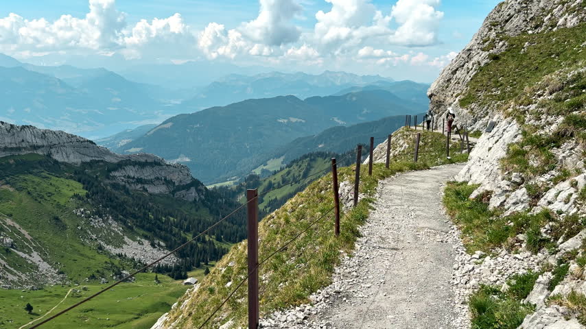 Narrow mountain trail crosses a steep rocky slope on Mount Pilatus outdoors, Elevated hiking route overlooks green valleys and distant Swiss peaks