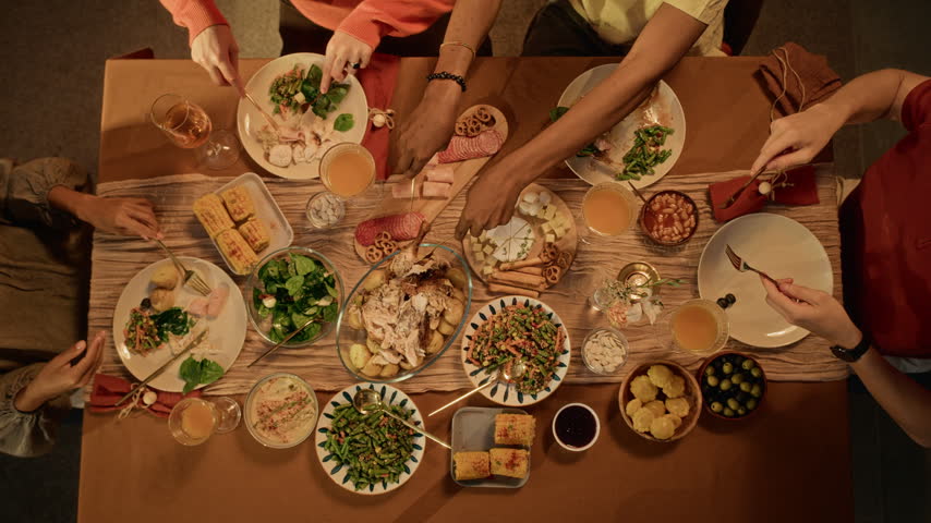 Multiethnic friends sharing food and eating festive meal during Friendsgiving dinner, top view. Ideal for Thanksgiving marketing, seasonal food campaigns, and holiday advertising