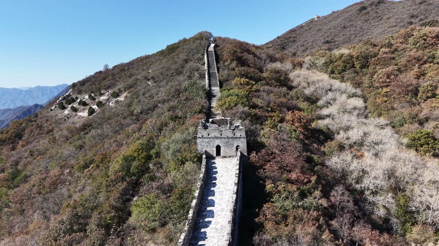 Static aerial looking down at a Great Wall watchtower with the wall climbing steeply uphill behind it, autumn foliage covering the surrounding mountain slopes, Huairou District, Beijing, China.