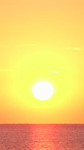 The sun sets over the ocean, casting warm orange and yellow hues. Gentle waves reflect the colorful sky, creating a serene atmosphere along the coastline.