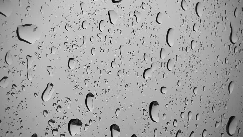Sirvintos, Lithuania - September 10 2026: A close-up full-frame macro capturing raindrops clinging to a smooth glass surface. The image displays a high density of water droplets, varying from small fine beads to larger, elongated droplets sliding down the transparent pane. Soft grey and silver light filters through the glass, creating dark curved outlines, bright highlights, and smooth refractive reflections within each individual drop. The visual composition emphasizes texture, surface tension, fluid dynamics, and wet weather ambiance. Visual themes focus on rainy days, atmospheric macro video, water droplets, glass textures, abstract moisture patterns, and calm moody weather conditions. This high-resolution stock photograph is ideal for weather forecasts, abstract backdrops, product design overlays, nature media, and commercial stock photo collections.